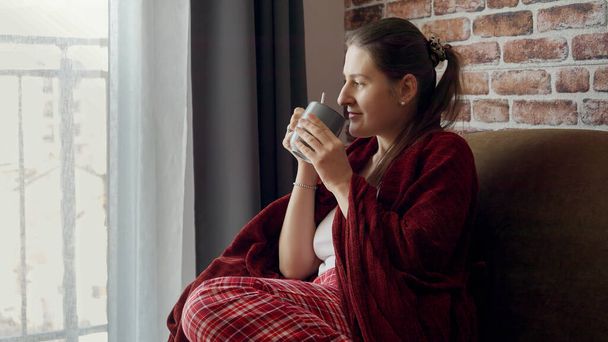 Krásná elegantní žena sedí na pohovce, pije kávu a dívá se z okna. Žena odpočívající doma, životní styl a relaxace - Fotografie, Obrázek