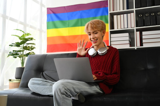 Delighted fiatal meleg férfi használ laptop kanapén a nappaliban LMBT büszkeség szivárvány zászló a háttérben. - Fotó, kép