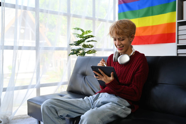 Positieve tiener gay man in rood trui met behulp van digitale tablet in heldere woonkamer met regenboog trots vlag op de achtergrond. - Foto, afbeelding