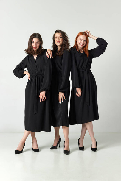 Full-length portrait of smiling, happy, beautiful young women in black dresses against light studio background. Concept of elegant fashion, female beauty, business, shopping, sales, ad - Φωτογραφία, εικόνα