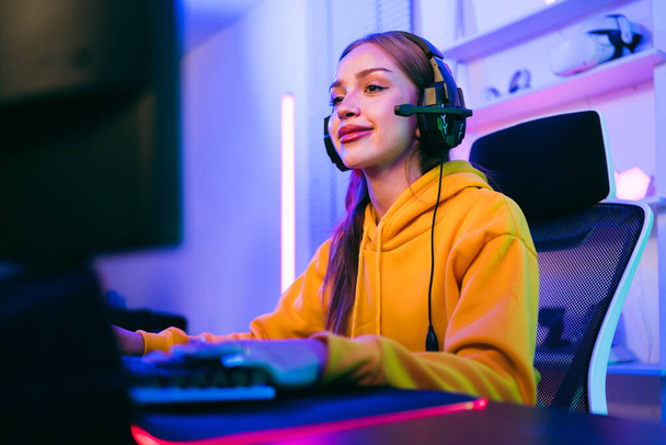 Young Caucasian woman professional gamer wearing yellow hoodie sits on a chair with a gaming table with pc, keyboard, monitor, microphone. Prepare for competition, cast gameplay or record a podcast. - Foto, afbeelding