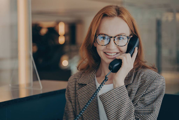 Smiling redhead office worker in plaid jacket and glasses, discussing project completion date on landline phone, office background blurred. - Photo, image