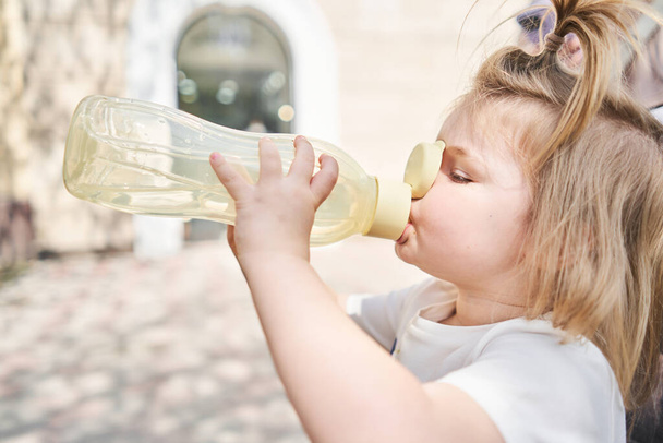 uma menina encantadora bebe água de uma garrafa de esportes em um parque da cidade. um retrato de close-up de uma criança que bebe de uma garrafa em um dia quente de verão. Foto de alta qualidade - Foto, Imagem