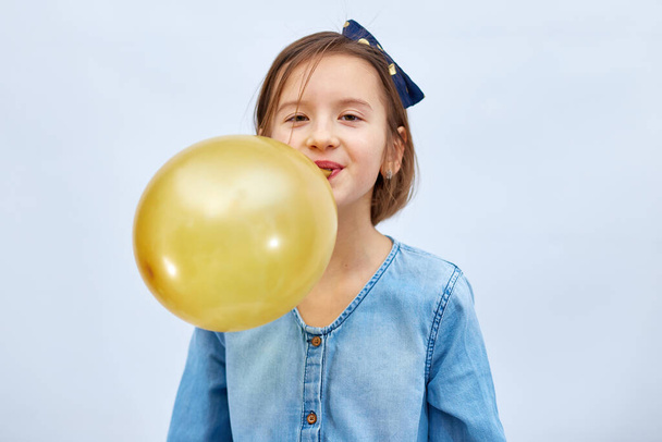 Pretty little girl in casual denim dress blowing, inflate yellow balloon on white background, studio - Fotó, kép