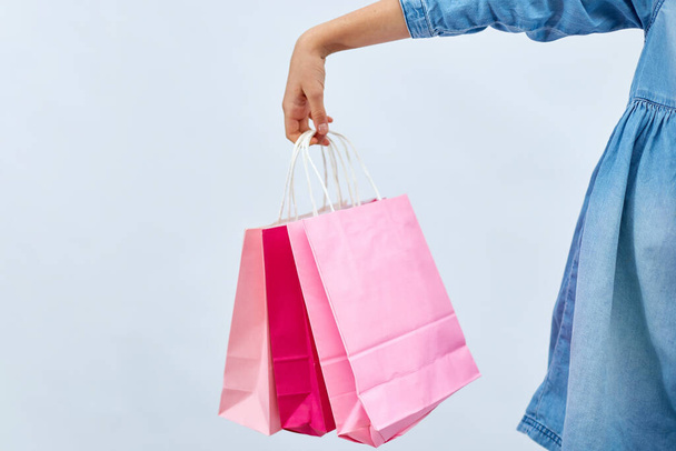 Unrecognizable child girl in casual denim dress hold in hand pink shopping bags on white background, studio, Shopaholic shopping and fashion, discount sale concept, copy space, banner. Black friday - 写真・画像