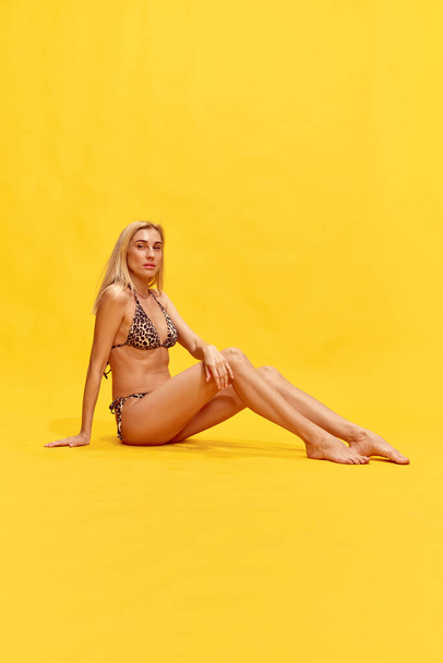 Beautiful blonde young woman with slim, fit body sitting, posing in stylish swimsuit against yellow studio background. Concept of summertime, fashion, female beauty, vacation, lifestyle, ad - Photo, Image