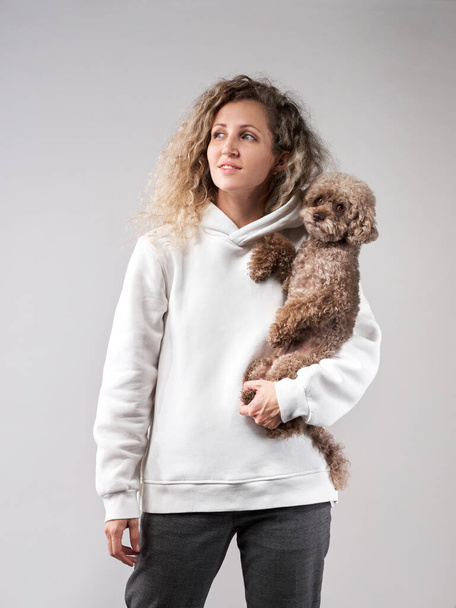 счастливая девушка с собакой в фотостудии на сером. Кудрявая женщина в белой толстовке с шоколадным пуделем на руках. - Фото, изображение