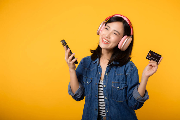 デニムの服を着た笑顔とスマートフォンやクレジットカードを持つヘッドフォンを身に着けて幸せな成功を収めた肖像アジアの若い女性。ショッピングオンライン携帯電話エンターテイメントライフスタイルコンセプト. - 写真・画像