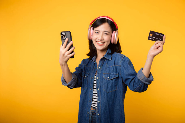 Asyalı genç bir kadının portresi mutlu bir gülümsemeyle kot elbise ve kulaklık giyerek elinde akıllı telefon ve kredi kartıyla. Çevrimiçi cep telefonu eğlence yaşam tarzı konsepti alışverişi. - Fotoğraf, Görsel