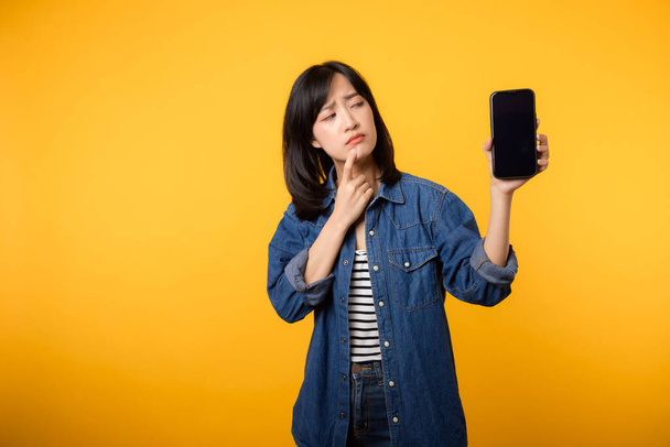 Портрет красивой молодой азиатской женщины с сомнением одеты в джинсовый пиджак, показывающий экран смартфона изолировать на желтом фоне студии. Новая концепция приложений для смартфонов - Фото, изображение