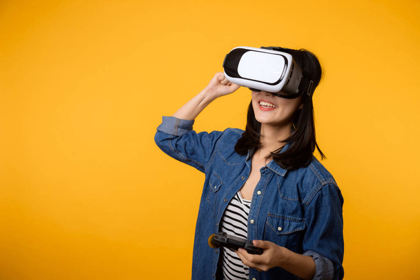 Asian young woman happy success wearing virtual reality headset game entertainment and holding joystick controller isolated on yellow background. Winner of cyber future video game concept. - Fotoğraf, Görsel