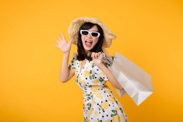 Muotokuva houkutteleva onnellinen nuori aasialainen nainen trendikäs kevät mekko, hattu, aurinkolasit muoti ja paperipussi eristetty keltaisella taustalla. Kesän myynti ostoksia käsite. - Valokuva, kuva
