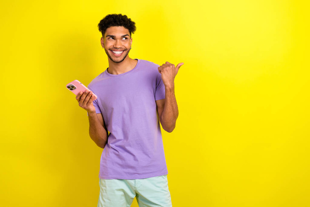 Photo of handsome blogger guy hold smart phone look indicate finger empty space isolated on yellow color background. - 写真・画像