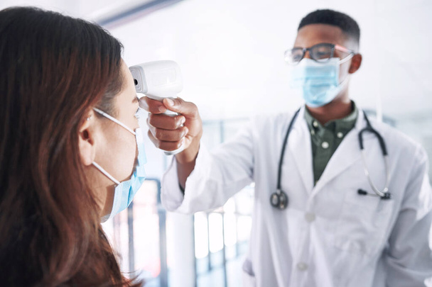 A lázmérés kötelező. egy fiatal orvos maszkot visel és egy beteggel áll a klinikán, miközben a lázát méri. - Fotó, kép