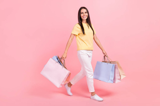 Full length photo of funky shiny woman dressed yellow t-shirt walking holding shoppers isolated pink color background. - Photo, Image