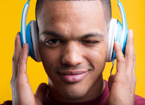 Le jeune homme fait un clin d'œil en écoutant passionnément de la musique ou du podcast, tenant des écouteurs bleus, sur fond jaune. Cheveux courts rasés, peau marron foncé, t-shirt bordeaux - Photo, image