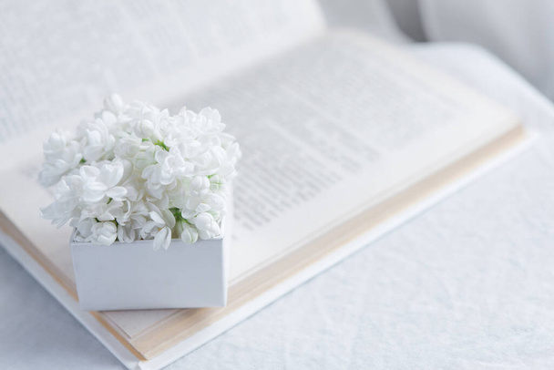 Close-up of a bouquet of white flowers in a white gift box on an open book on a white textile background. Slow life concept. Greeting card mockup - Фото, изображение