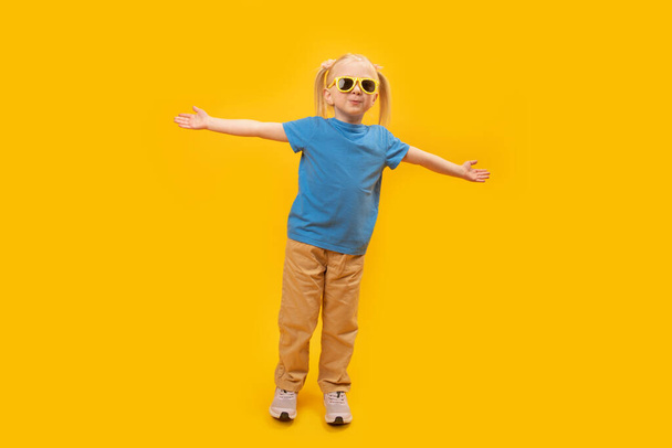 Sarı pantolonlu, mavi tişörtlü, şık güneş gözlüklü şık küçük sarışın kız. Çocuk sarı arka planda el kaldırdı ve kameraya baktı. - Fotoğraf, Görsel