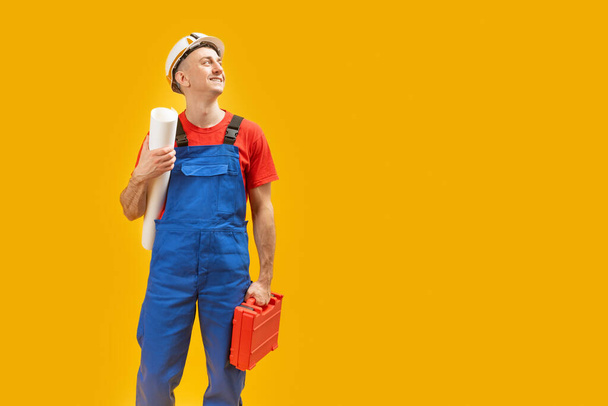 Jonge ingenieur met helm met blauwdrukken en gereedschapskist die wegkijkt over geïsoleerde gele achtergrond. Kopieer ruimte, mockup. - Foto, afbeelding