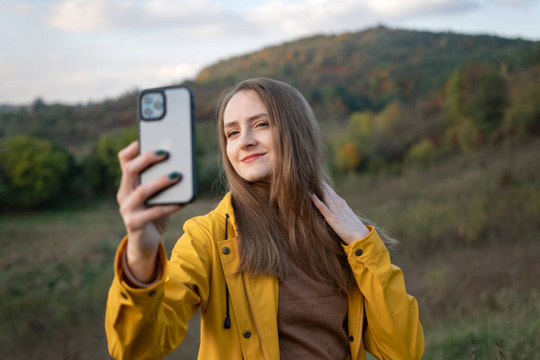 Akıllı telefonlu genç bir kadının portresi, arka planda dağ. Sonbahar doğasının sarı ceketli kızı.. - Fotoğraf, Görsel
