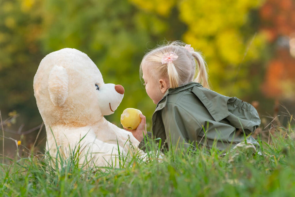 Little girl with two tails plays with teddy bear on grass near autumn forest. Fair-haired child in nature with soft toy. - Фото, зображення