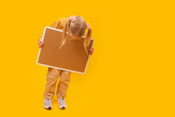 Little girl holding empty board and looking down. Yellow background. Space for your text. Copy space, mockup. - Photo, Image