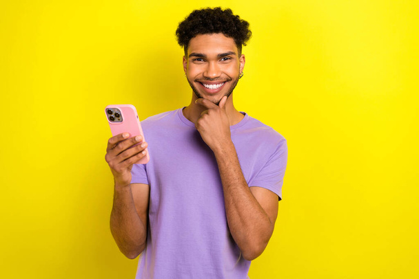 Photo of cheerful minded creative person hand touch chin toothy smile hold smart phone isolated on yellow color background. - Fotó, kép