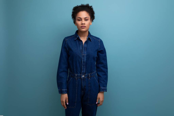 portrait of a young woman with dark skin and black curly hair dressed in blue denim overalls isolated background with copy space. - Photo, Image