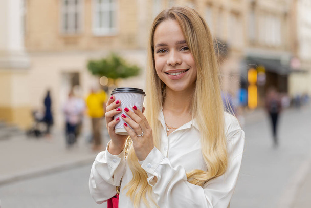 Fröhliche blonde junge Frau genießt den morgendlichen Kaffee und lächelt im Freien. Entspannen, Pause machen. Teenager-Mädchen, das in der städtischen Straße spaziert und Kaffee to go trinkt. Stadtleben im Freien - Foto, Bild
