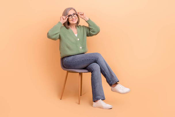 Full body length cadre of satisfied senior businesswoman wear khaki shirt touch eyeglasses sit chair isolated on beige color background. - Fotoğraf, Görsel