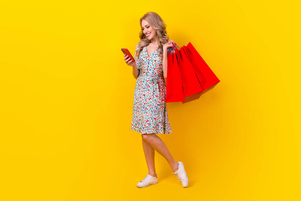 Full body photo of young blonde wavy hair girl hold bags shoppers clothing read phone virtual store isolated on yellow color background. - Foto, imagen