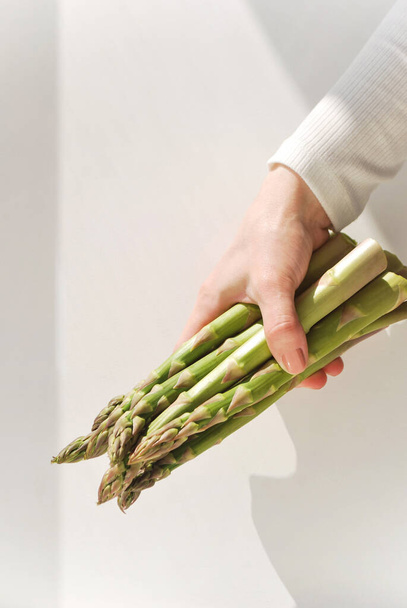 A women's hand holding green fresh asparagus on the background of a light wall with daylight and shadows. Green living and eco-friendly products - Foto, afbeelding