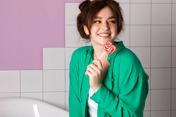 attractive beautiful girl stylish with bright makeup and double bun hair, emotional, facial expression, posing in bathroom, eating heart shaped lollipop, clean minimalist style, white tiles background - Photo, image