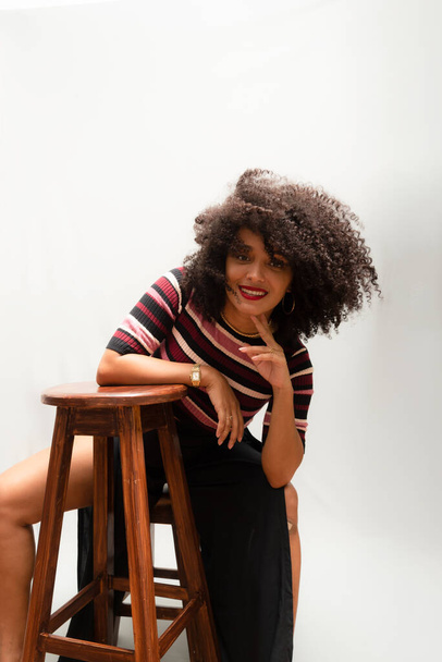 Portrait of a beautiful smiling cheerful young woman with curly hair sitting on a wooden stool. Isolated on light gray background. - Photo, Image