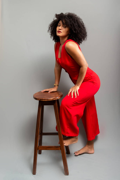 Young woman, standing, wearing a red outfit with curly hair posing next to a wooden stool. Isolated on light gray background. - Foto, Bild