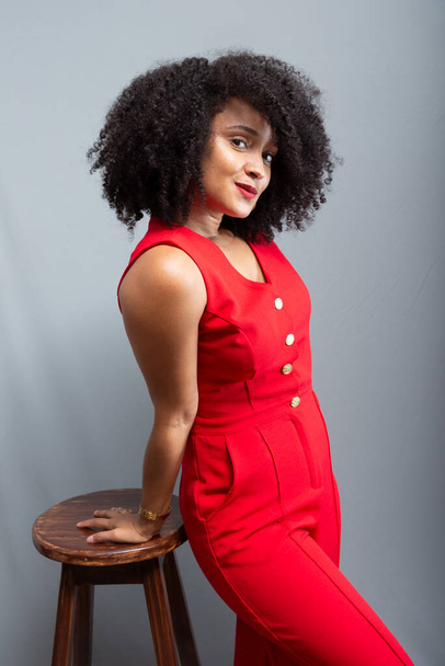 Młoda kobieta, stojąca w czerwonym stroju z kręconymi włosami, pozująca obok drewnianego stolca. Izolacja na jasnoszarym tle. - Zdjęcie, obraz