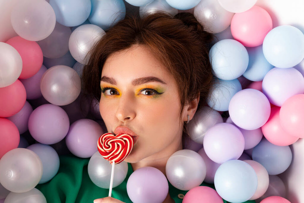 portrait face shot, attractive beautiful girl stylish with bright makeup and double bun hair, emotional, facial expression, lying in a pool filled with pastel multi colored plastic balls, top view. - Photo, Image