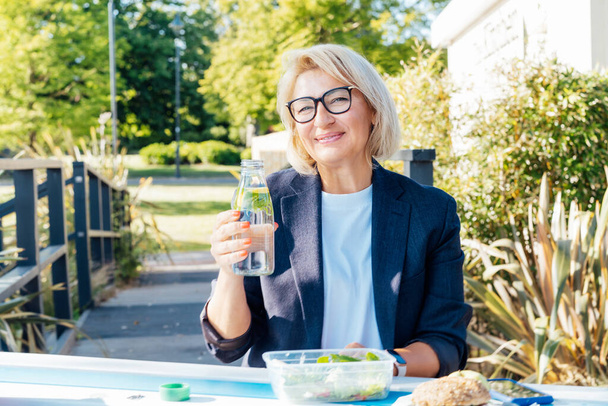 Healthy habit to drink water. Smiling middle aged business woman with bottle of water with lemon and mint during her outdoor lunch break. Control body hydration, Tracker water balance. Healthy living - Photo, Image
