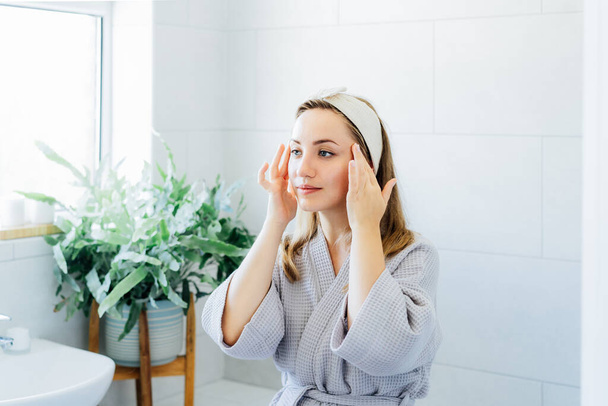 Young woman in bathrobe looking in mirror and doing manual self massage of face in the bathroom. Natural procedures for skin care. Home Beauty treatment, face gymnastics for youth. Selective focus. - Photo, Image