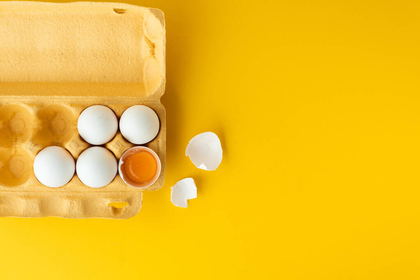 Flache weiße Eier in der braunen Schachtel auf gelbem Hintergrund. Draufsicht auf zerbrochenes Ei mit weißer Schale. Kopierplatz für einen freien Text. - Foto, Bild