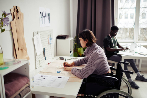 Criativa jovem mulher sentada em cadeira de rodas em sua mesa em estúdio e desenho padrão de costura - Foto, Imagem