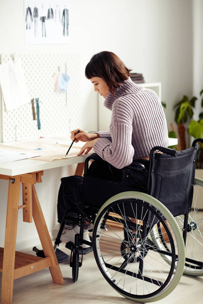 Tekerlekli sandalyedeki yaratıcı genç moda tasarımcısı proje için yaptığı dikiş düzenini kontrol ediyor. - Fotoğraf, Görsel