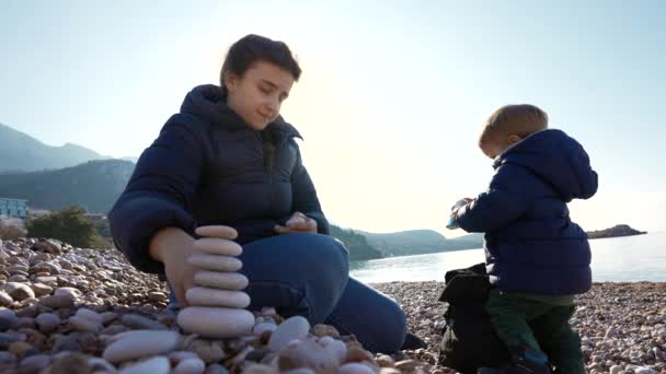 Madre e figlio costruiscono una torre di ciottoli su una spiaggia di pietra serena. Il filmato soleggiato e al rallentatore cattura la gioia e la concentrazione del loro sforzo collaborativo, creando un bellissimo ricordo in riva al mare - Filmati, video