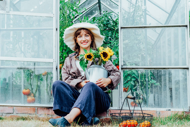 Portrait of smiling young female farmer woman holding watering can with fresh sunflowers bouquet, sitting near greenhouse. Urban farming lifestyle. Growing organic vegetables in garden - Photo, Image