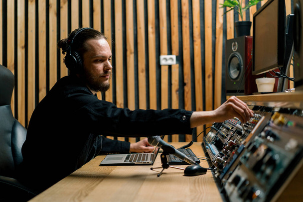 молодой звукорежиссер с наушниками, работающий в музыкальной студии с мониторами и эквалайзером, микширующим треки - Фото, изображение