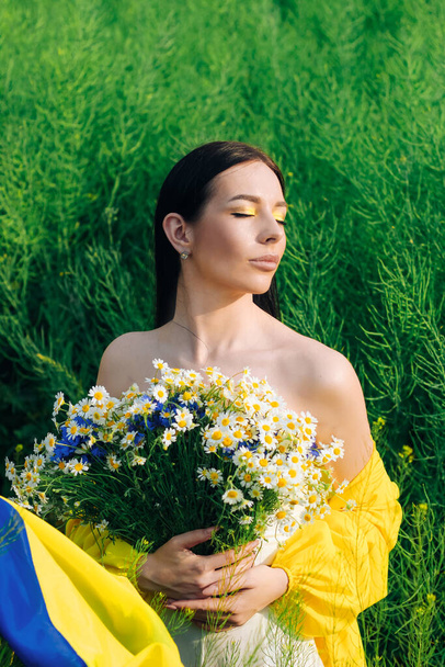 Українська дівчина з синьо-жовтим прапором в руках і букетом квітів на задньому плані фіолету. - Фото, зображення