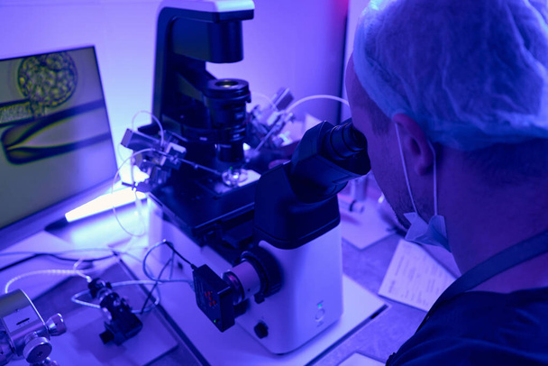 Reproductology laboratory worker looking in ocular lenses of microscope and working with micromanipulator, making injections to oocyte - Photo, image