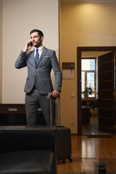 Hotel guest in a business suit communicates on a mobile phone, a man has a travel suitcase - Foto, Bild
