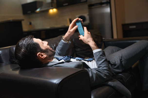 Hotel guest is relaxing on a leather sofa in the room, he is using a mobile phone - 写真・画像