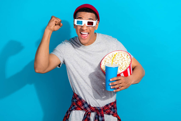 Portrait of crazy funky handsome guy raise fist hold soda drink popcorn watch movie 3d glasses isolated on blue color background. - 写真・画像